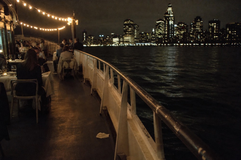 A dinner table set on a boat deck with the city skyline beyond, no text, no logos, no people